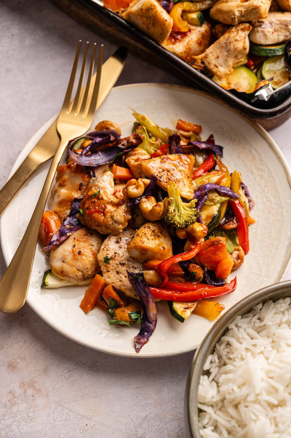 A plate of colorful stir-fried chicken with vibrant vegetables like red peppers, broccoli, and purple cabbage. A fork and knife are beside it, with a bowl of white rice nearby.