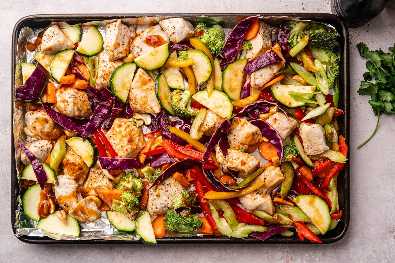 A colorful sheet pan loaded with seasoned chicken pieces, vibrant slices of zucchini, red cabbage, carrots, bell peppers, and broccoli, placed on foil.