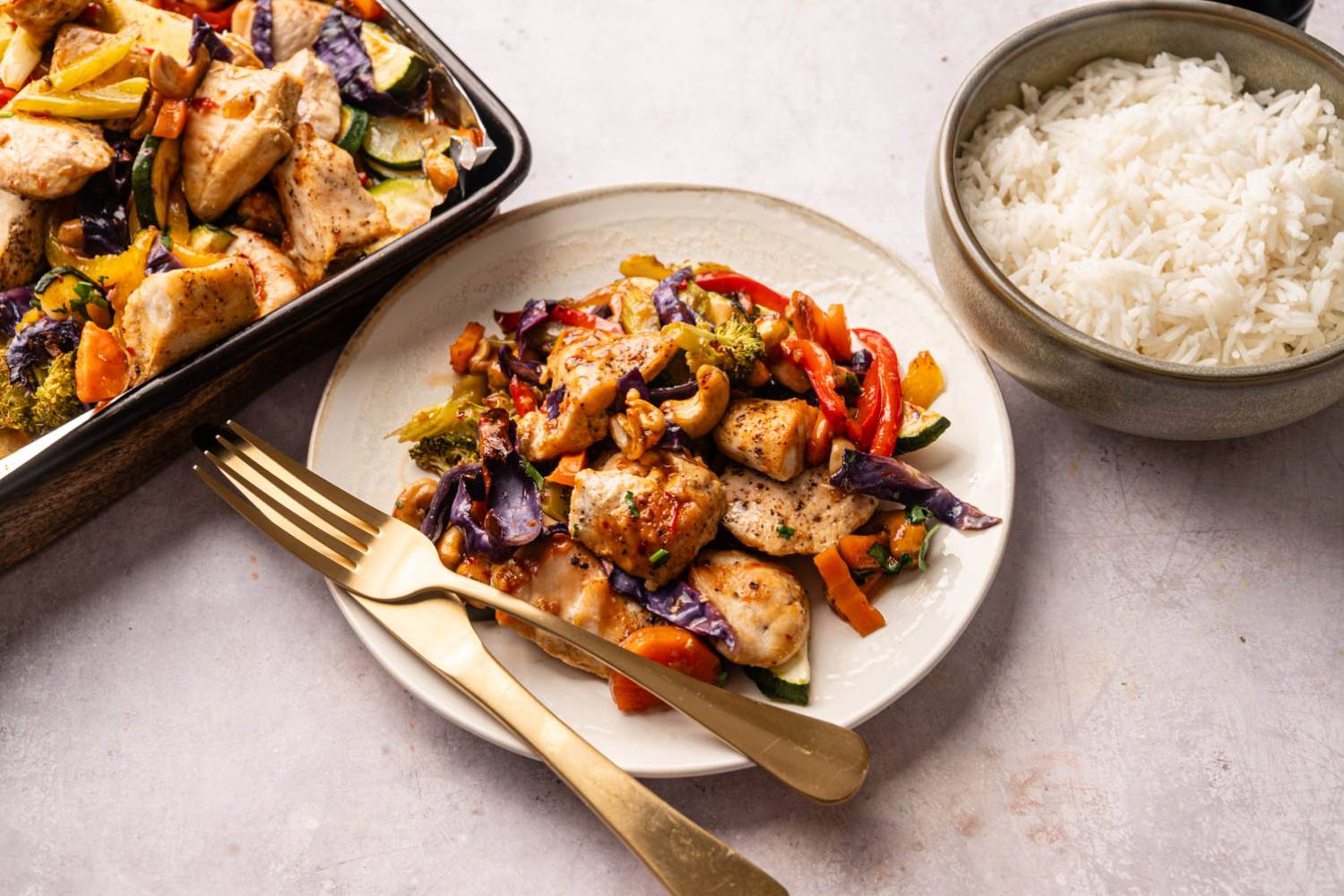 Sheet Pan Thai Cashew Chicken and Vegetables Stir-fried chicken with colorful vegetables, including red peppers and purple cabbage, on a white plate next to a bowl of white rice.