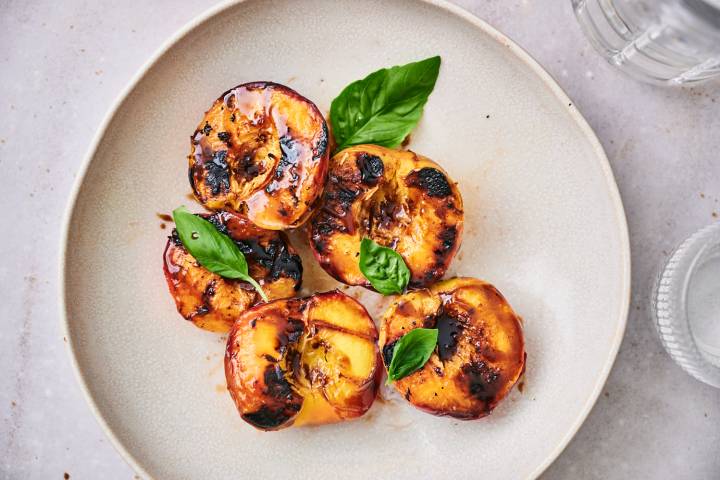 Grilled Peaches Grilled peaches with dark brown grill marks served on a plate with honey and fresh basil.