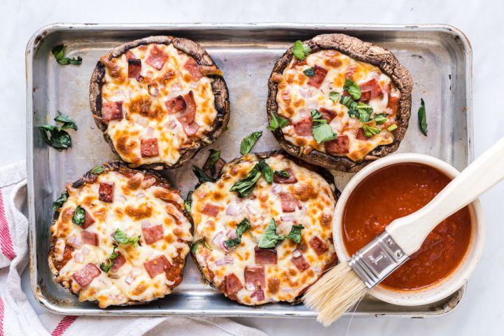 Portobello Mushroom Pizzas Portobello mushroom pizzas on a baking sheet with marinara sauce, melted cheese, basil, ham, and onions.