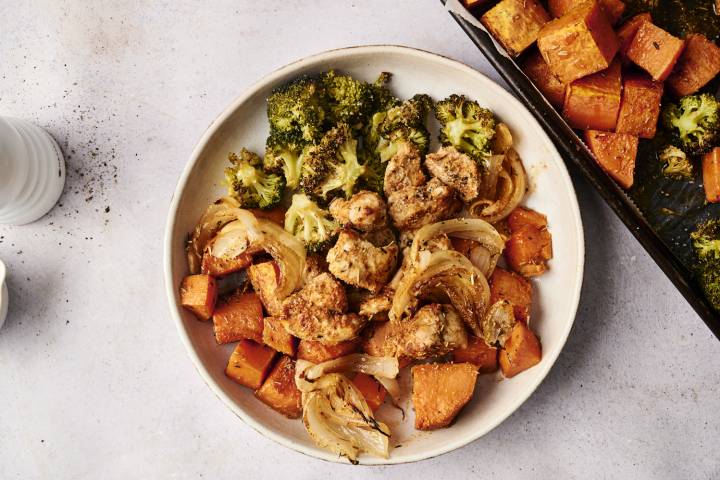 Sheet Pan Herb Chicken And Sweet Potatoes A serving of sheet pan herb chicken and sweet potatoes plated with roasted broccoli, caramelized onions, and seasoned sweet potato chunks. The plate is set on a light background with a portion of the sheet pan visible beside it.