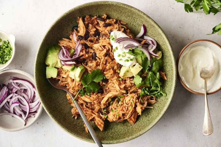 Slow Cooker Chicken Burrito Bowls A chicken burrito bowl topped with avocado, cilantro, red onions, sour cream, and chopped chives, served in a green ceramic dish with a spoon.