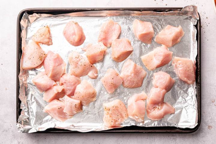 Sheet Pan Thai Cashew Chicken and Vegetables Chunks of raw chicken seasoned with black pepper scattered on a foil-lined baking tray.