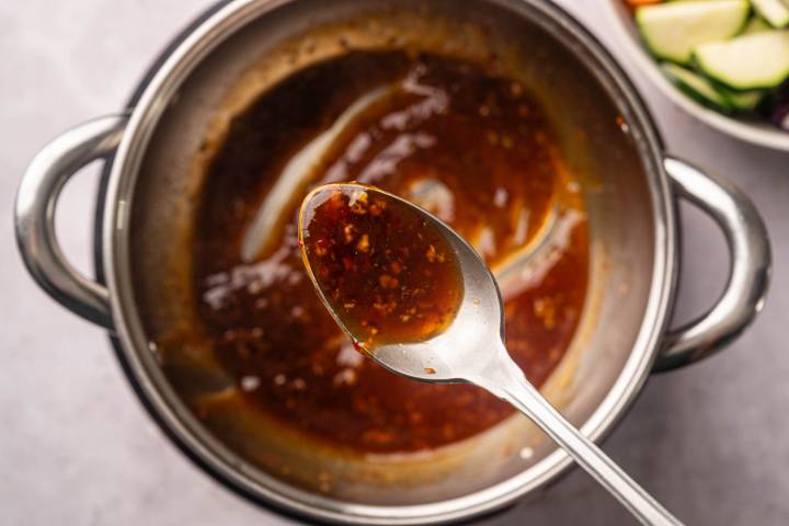 Sheet Pan Thai Cashew Chicken and Vegetables Close-up of a spoon holding a thick, dark red sauce with flecks of seasoning over a metal pot.
