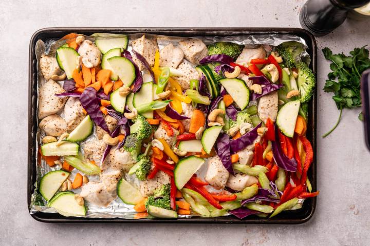 Sheet Pan Thai Cashew Chicken and Vegetables Sheet pan with baked chicken, zucchini, red bell peppers, broccoli, carrots, and cashews, arranged colorfully. Fresh cilantro on the side.