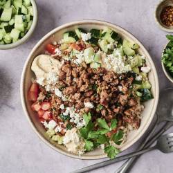 Chicken Kofta Bowls Bowl of hummus topped with seasoned ground meat, cucumber, tomatoes, feta, and fresh parsley, surrounded by ingredient bowls and utensils.