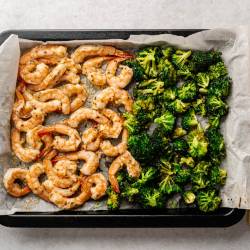 Garlic Parmesan Roasted Shrimp and Broccoli Roasted shrimp and broccoli on a baking sheet lined with parchment paper. Olive oil, lemon wedges, and a pepper mill are nearby. Bright and appetizing.