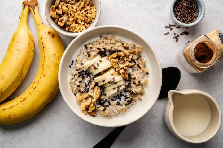 Banana Protein Oatmeal A creamy oatmeal bowl topped with banana slices, walnuts, and chocolate sprinkles. Nearby are ripe bananas, walnuts, chocolate sprinkles, and milk. Warm, inviting tone.