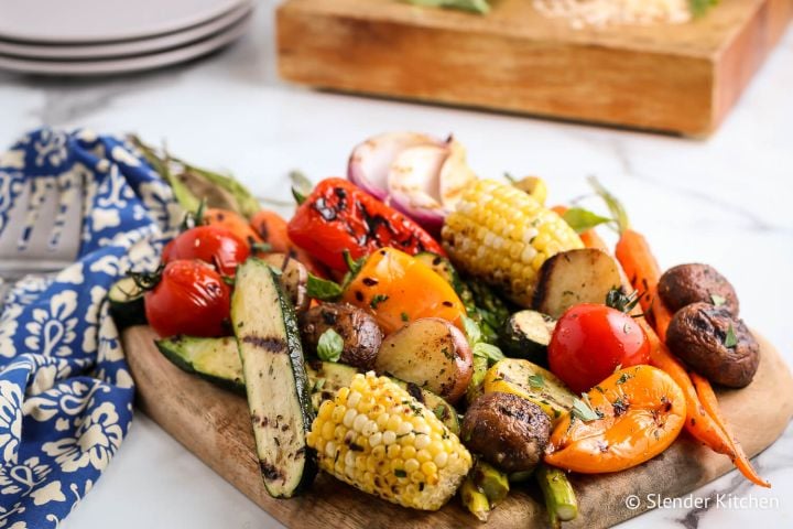 Easy Grilled Vegetables Grilled vegetables including zucchini, peppers, tomatoes, potatoes, and carrots on a wooden cutting board.