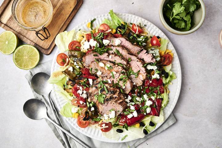 Grilled Steak Fajita Salad A vibrant salad topped with sliced grilled beef, assorted veggies, feta cheese, and cilantro, accompanied by lime halves and dressing.