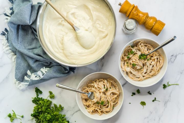 Cauliflower Alfredo Sauce Skinny Alfredo sauce in a large pan with a wooden spoon and served in two bowls with spaghetti and parsley.