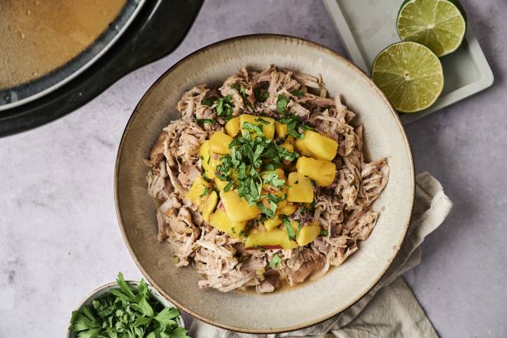 Slow Cooker Jerk Chicken A bowl of shredded meat topped with diced mango and fresh cilantro, surrounded by lime halves and fresh herbs.