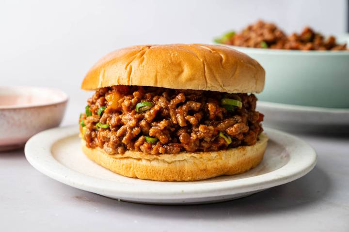 Three Ingredient Sloppy Joes A juicy sloppy joe sandwich is centered on a white plate. The bun is golden brown, holding a savory, saucy ground meat filling.