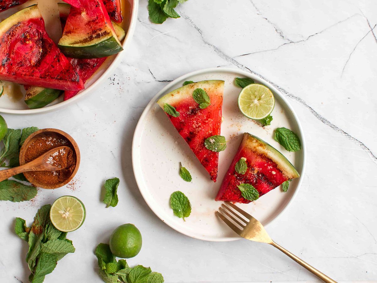 Grilled Fruit Slices of grilled watermelon on a white plate garnished with fresh basil, cut limes, and chili powder on the side.
