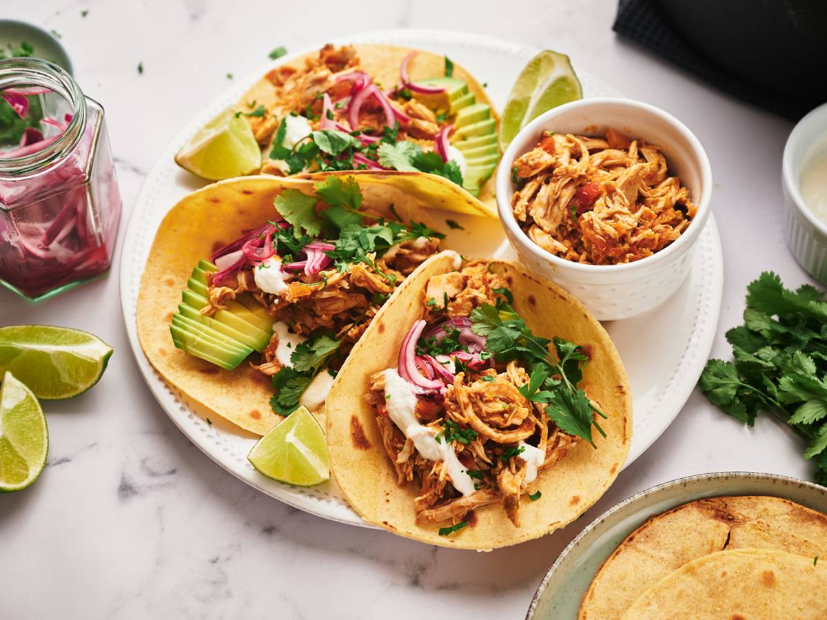 Chicken Tacos 3 Shredded chicken tacos with avocado, pickled onions, cilantro, and limes served on a round plate.