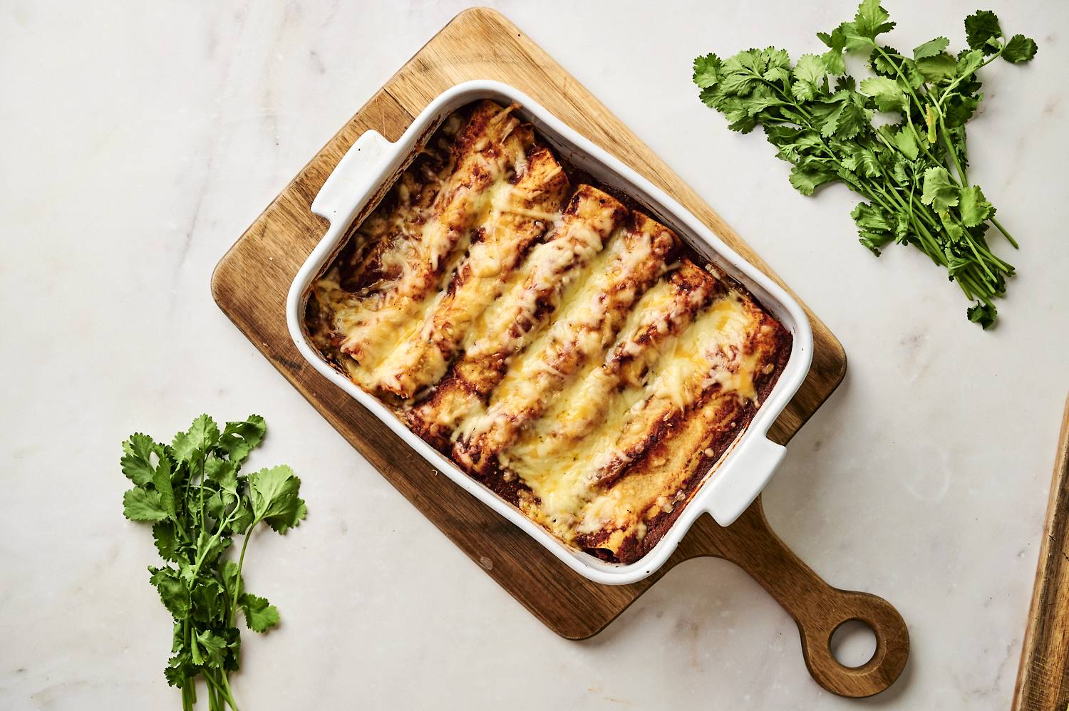 Cheesy black bean enchiladas in a white baking dish, topped with diced avocado and fresh cilantro, with a golden serving spoon lifting a portion.