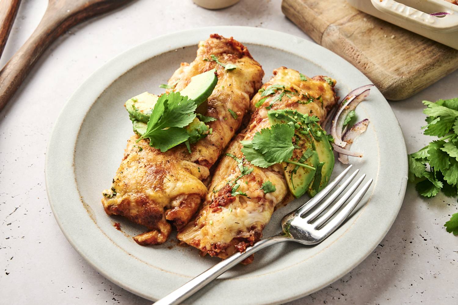 Two cheesy chicken enchiladas garnished with fresh cilantro and avocado slices on a plate, ready to enjoy.