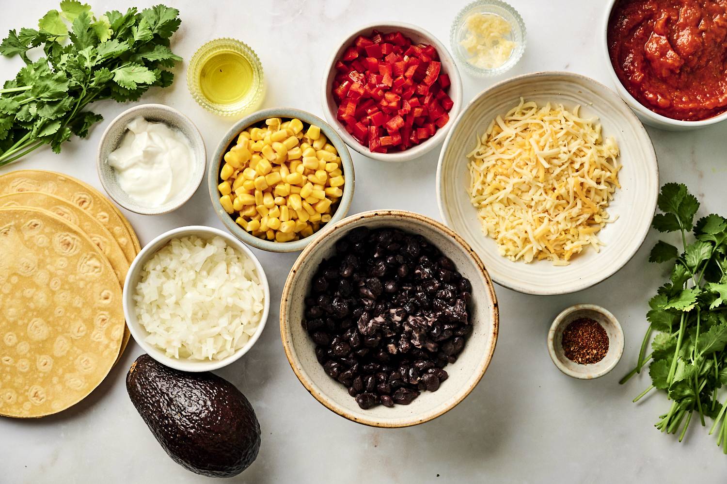 A flat lay of fresh ingredients for black bean enchiladas, including tortillas, black beans, corn, diced red peppers, shredded cheese, enchilada sauce, and seasonings, arranged on a white surface.