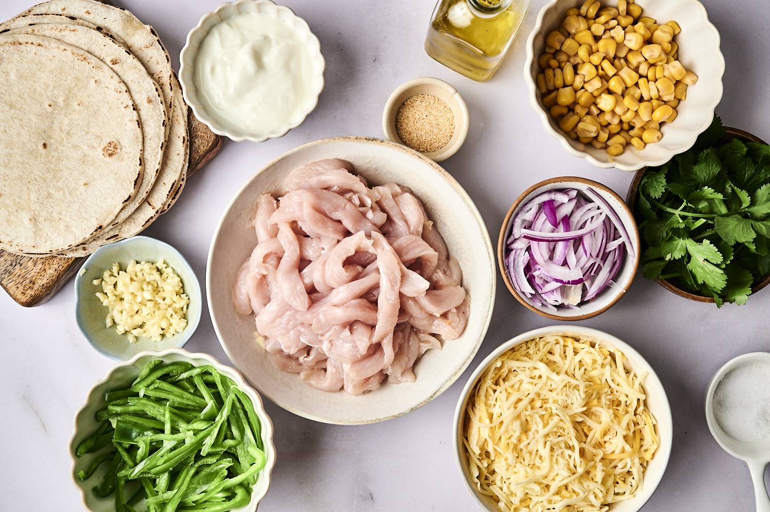 A flat lay of ingredients for cooking, including sliced chicken, tortillas, green peppers, onions, corn, cheese, garlic, cilantro, and yogurt.