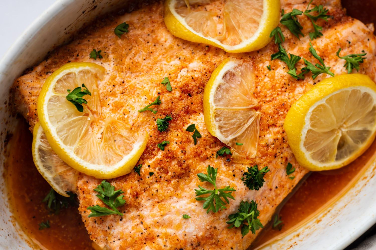 Salmon baked with lemon pepper seasoning, butter, and lemon slices in a white dish,