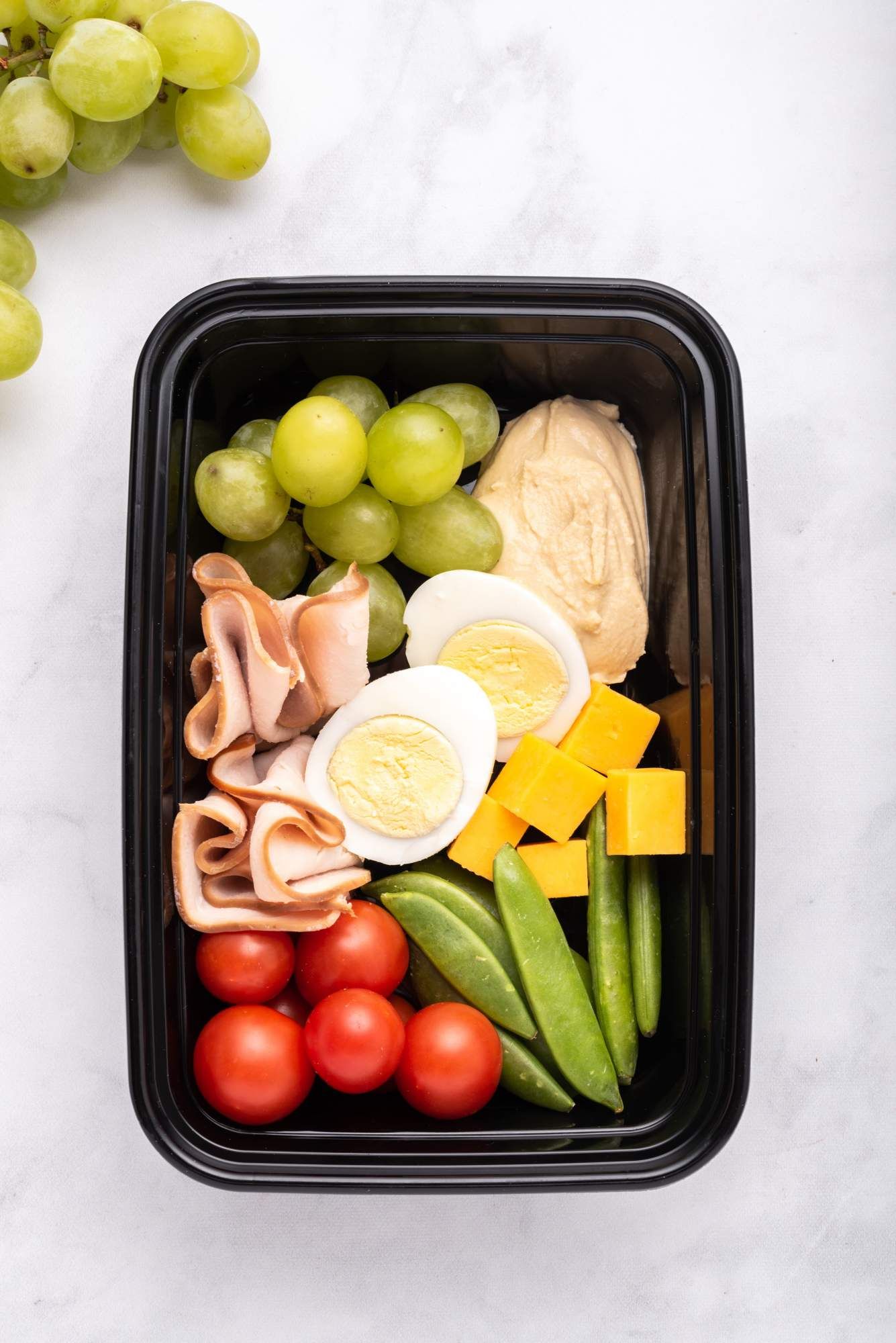 Snack box with protein filled snacks including hummus, hard boiled eggs, cheddar cheese, turkey, grapes, and vegetables.
