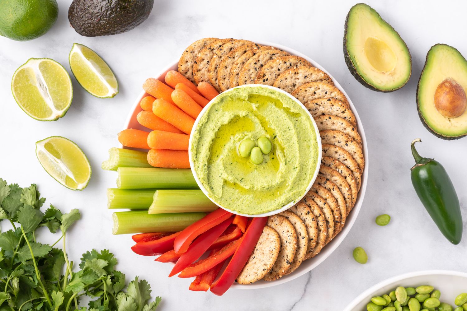 Edamame hummus with frozen edamame, cilantro, avocado, and lime on a plate with vegetables and crackers.