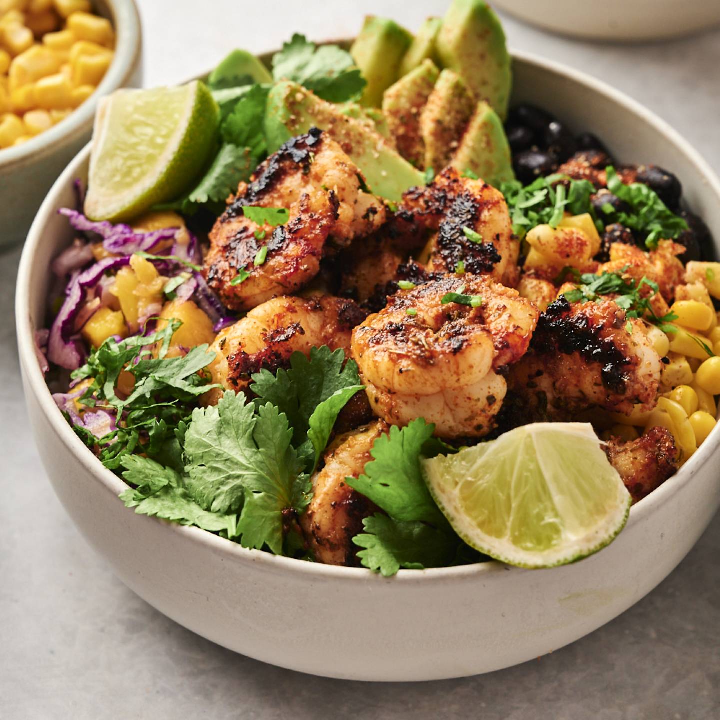 Blackened shrimp bowl topped with grilled shrimp, avocado slices, lime wedges, cilantro, and surrounded by corn, purple cabbage, and black beans.
