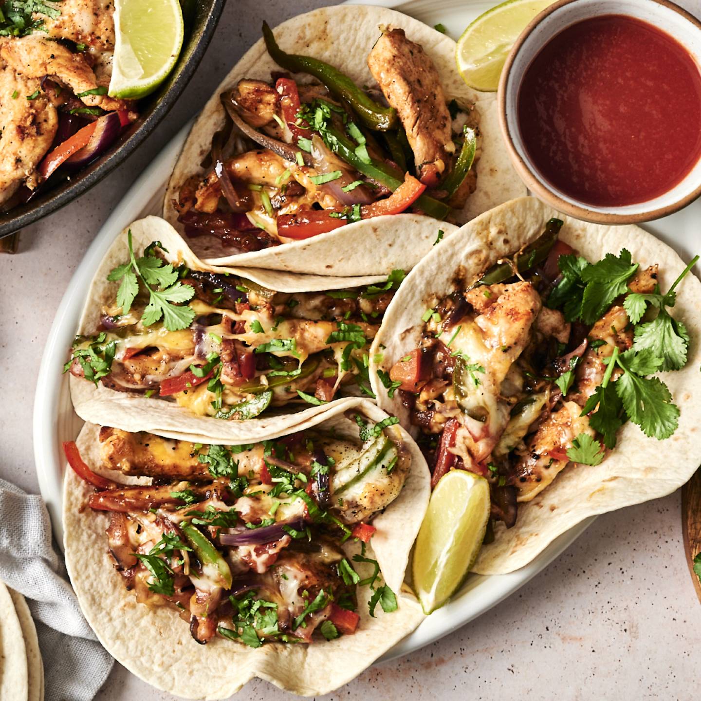 A plate of chicken alambre tacos topped with cilantro, melted cheese, and lime wedges, with a side of salsa, accompanied by a skillet of sautéed chicken and vegetables.