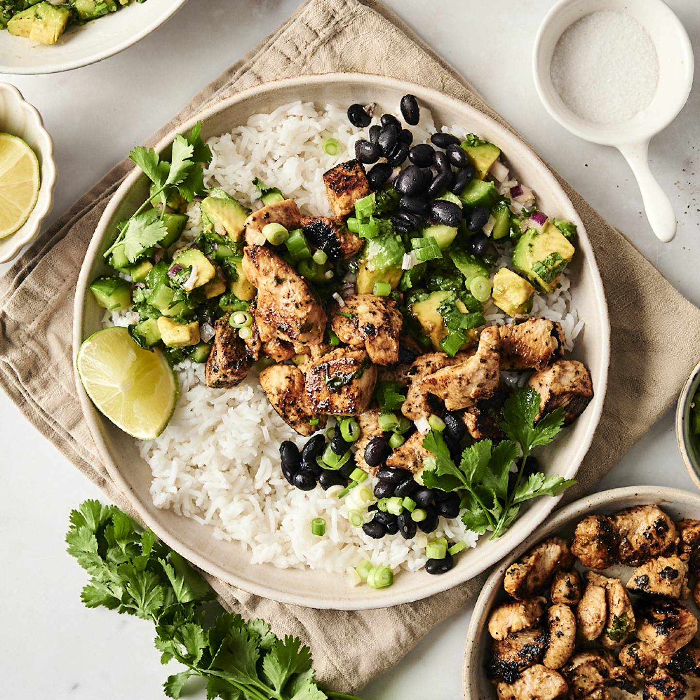 A bowl filled with grilled chili-lime chicken, white rice, black beans, avocado salsa, and fresh cilantro, garnished with lime wedges and green onions.