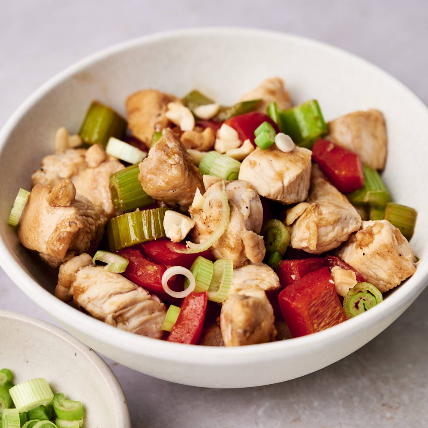 A bowl of Healthy Kung Pao Chicken featuring chunks of chicken, red bell peppers, celery, and garnished with green onions and peanuts.