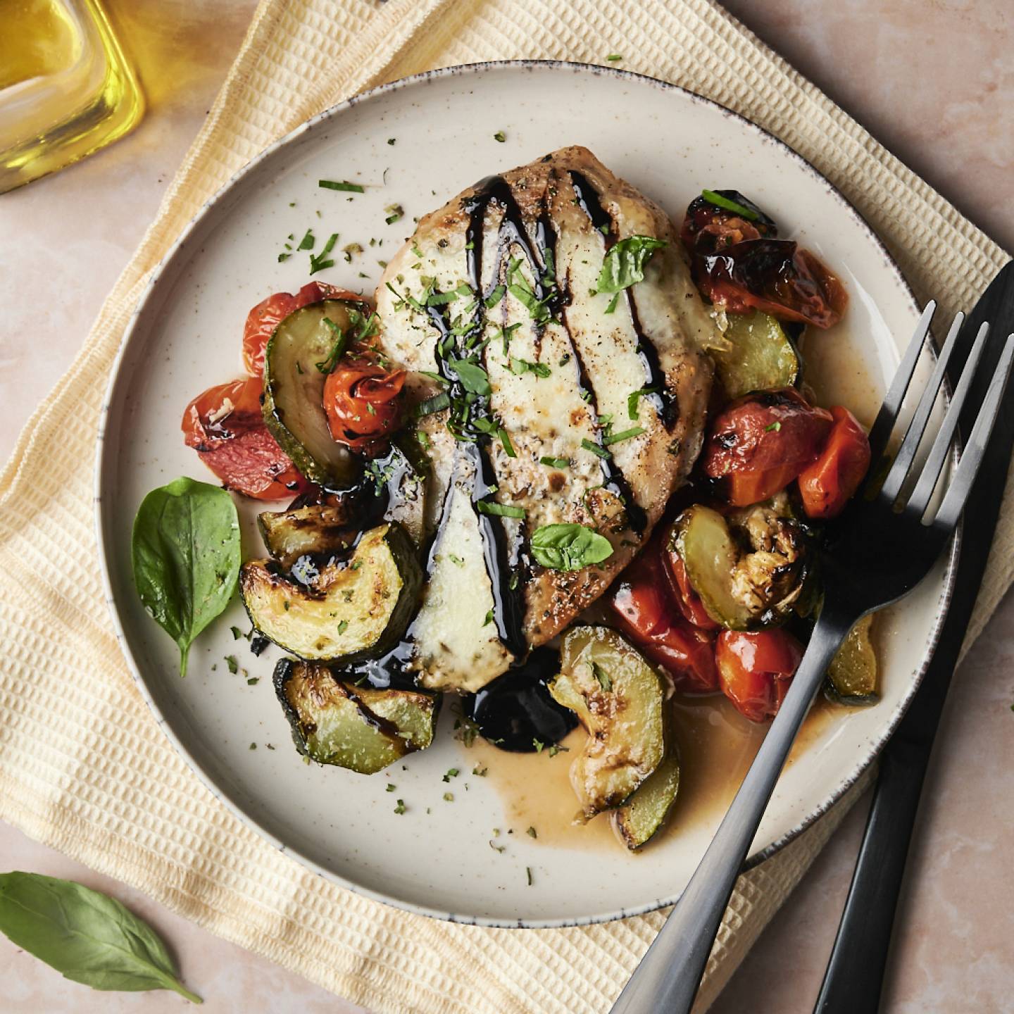 A plate of caprese chicken with roasted zucchini and tomatoes, topped with balsamic glaze and fresh basil.