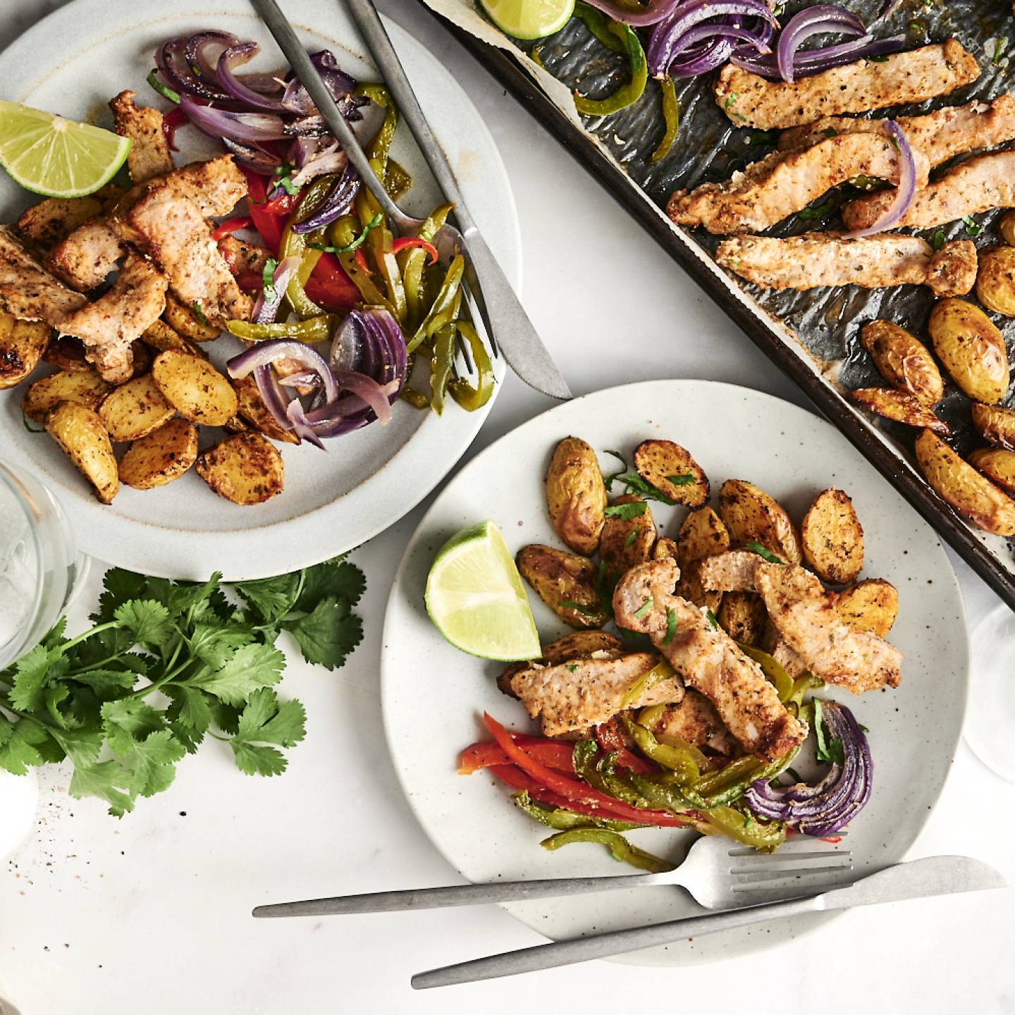 A sheet pan meal featuring seasoned pork chops, roasted potatoes, and sautéed bell peppers with red onions, served on plates with fresh cilantro and lime wedges.