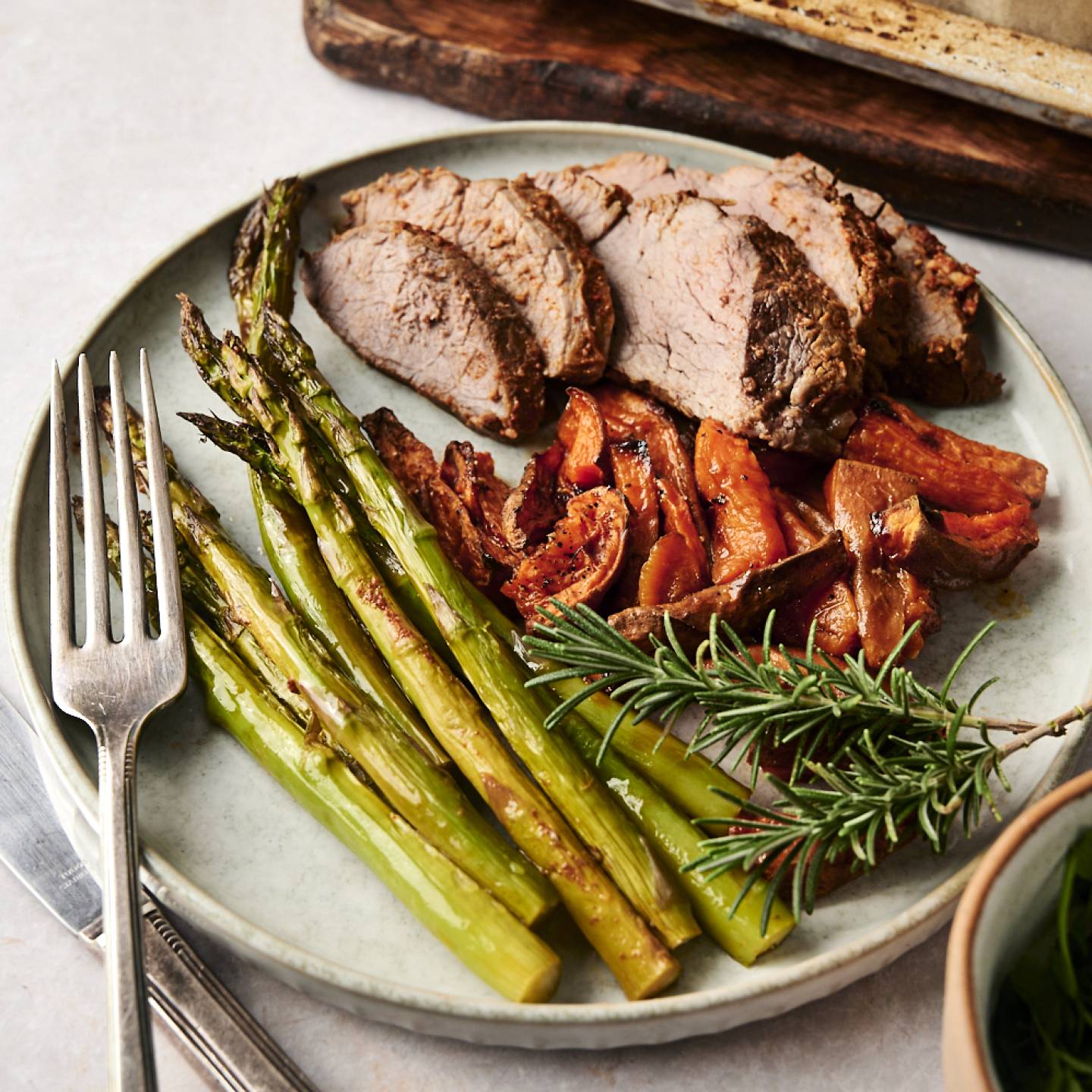 A close-up view of a dinner plate featuring juicy rosemary-seasoned steak slices, perfectly roasted asparagus, and golden sweet potato wedges, plated elegantly with a sprig of fresh rosemary and accompanied by a fork and knife