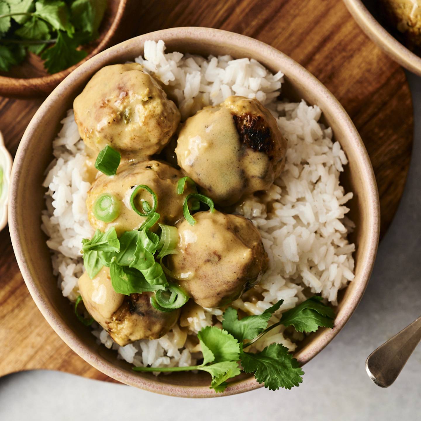 A bowl of white rice topped with Thai curry chicken meatballs, coated in a creamy sauce and garnished with fresh cilantro and sliced green onions.