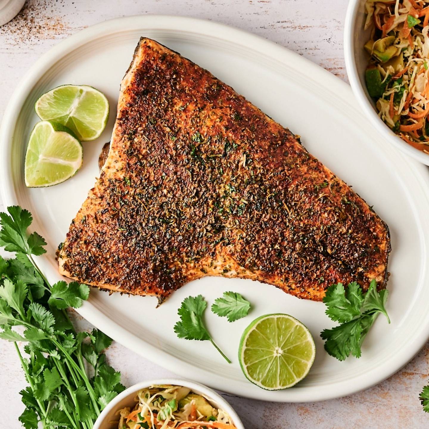 A whole taco spiced salmon fillet is served on a white platter with lime wedges, fresh cilantro, and bowls of slaw on the side.