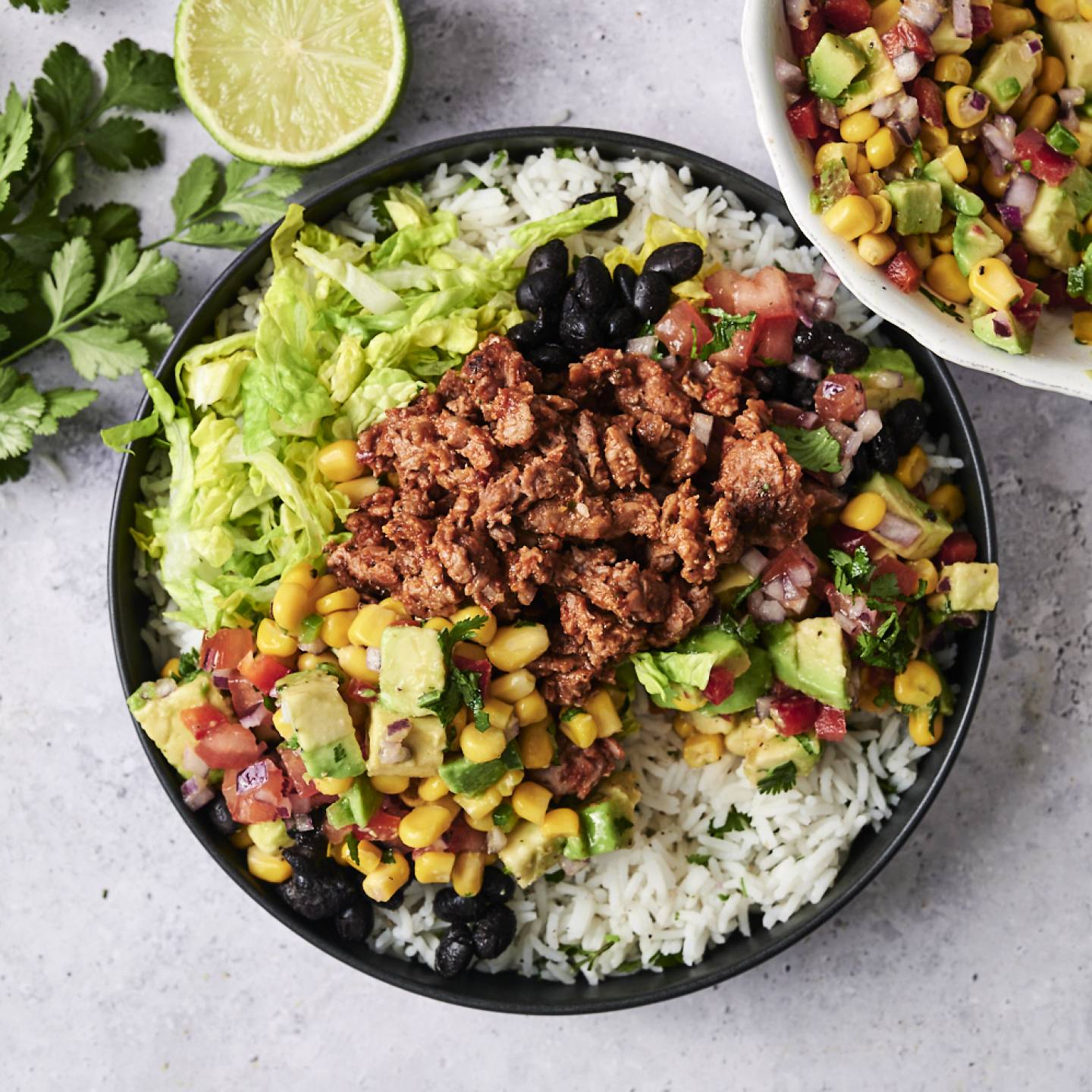 A vegan ground beef burrito bowl is served with cilantro lime rice, black beans, lettuce, seasoned plant-based ground beef, avocado, corn, and fresh pico de gallo, garnished with chopped cilantro and a lime wedge.
