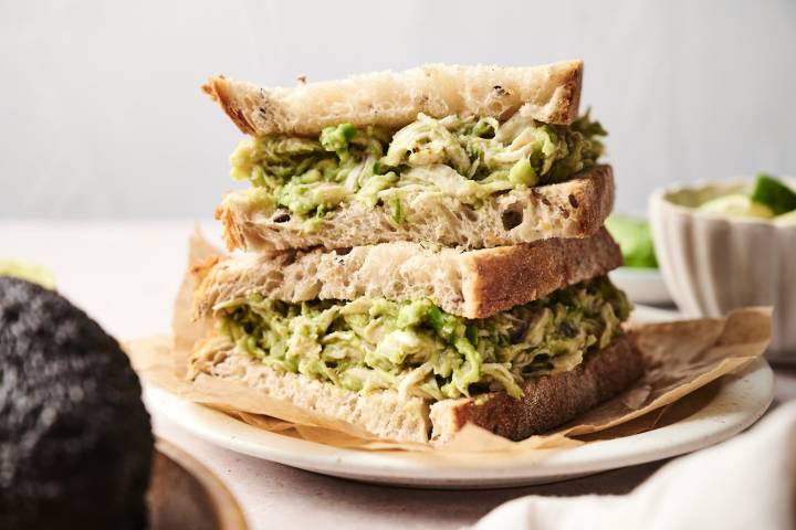 Close-up of a stacked sandwich made with thick slices of rustic bread and filled generously with creamy avocado chicken salad. The sandwich is served on a parchment-lined plate, ready to enjoy.