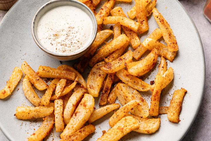 Crispy seasoned fries are arranged on a plate with a small bowl of creamy dipping sauce. The fries are golden and dusted with spices.