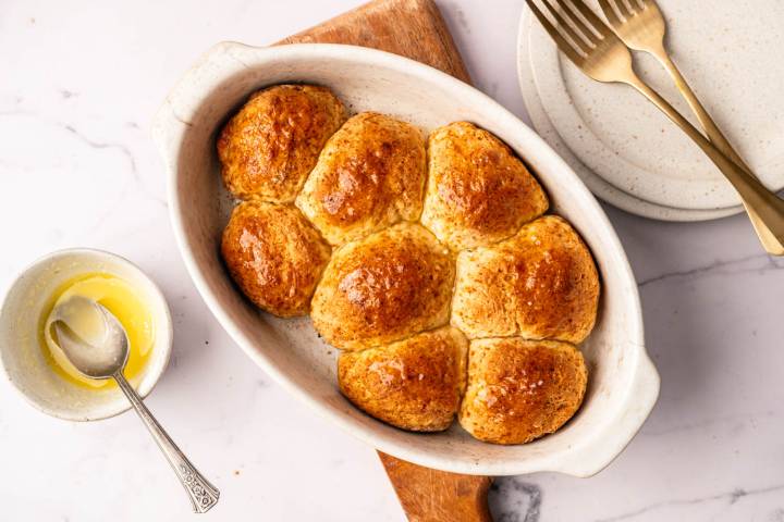 Golden brown dinner rolls in an oval dish on a wooden board. Nearby, a small bowl of melted butter with a spoon, and two forks on stacked plates. Warm, inviting tone.