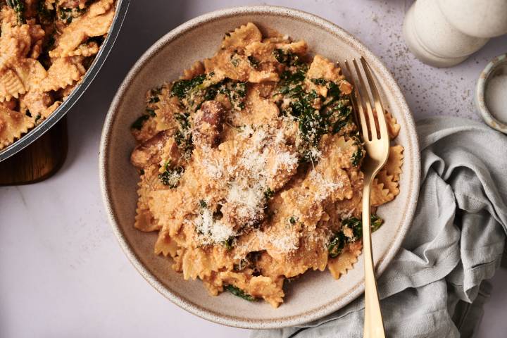 Creamy high-protein pasta made with bowtie noodles, sausage, spinach, and ricotta, served in a beige bowl and topped with grated Parmesan cheese. A gold fork rests on the side.  