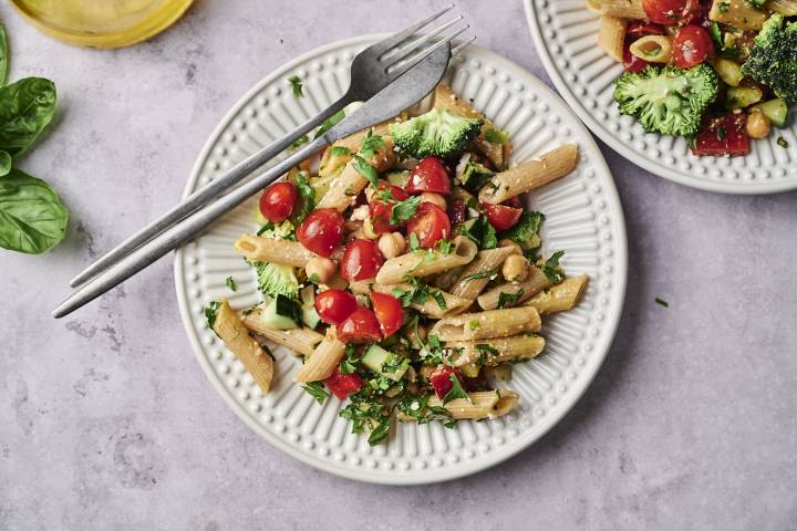 A vibrant plate of pasta mixed with cherry tomatoes, broccoli, and herbs, accompanied by a glass of olive oil and fresh basil leaves.