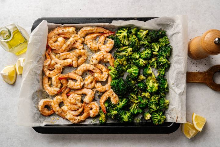Roasted shrimp and broccoli on a baking sheet lined with parchment paper. Olive oil, lemon wedges, and a pepper mill are nearby. Bright and appetizing.