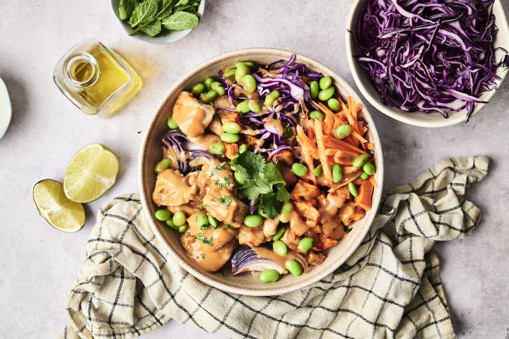 A vibrant bowl of chicken, edamame, purple cabbage, and carrots drizzled with sauce, surrounded by lime, mint, and olive oil.