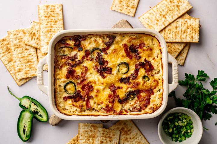 A creamy baked dish topped with bacon and jalapeños is centered in a rectangular dish. Surrounding the dish are several crackers, sliced jalapeño and fresh parsley, with a small bowl of chopped green onions.