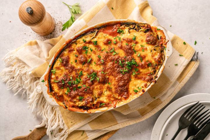 Baked lasagna in an oval dish on a checkered towel, garnished with chopped parsley. A pepper grinder and a plate with utensils are nearby.