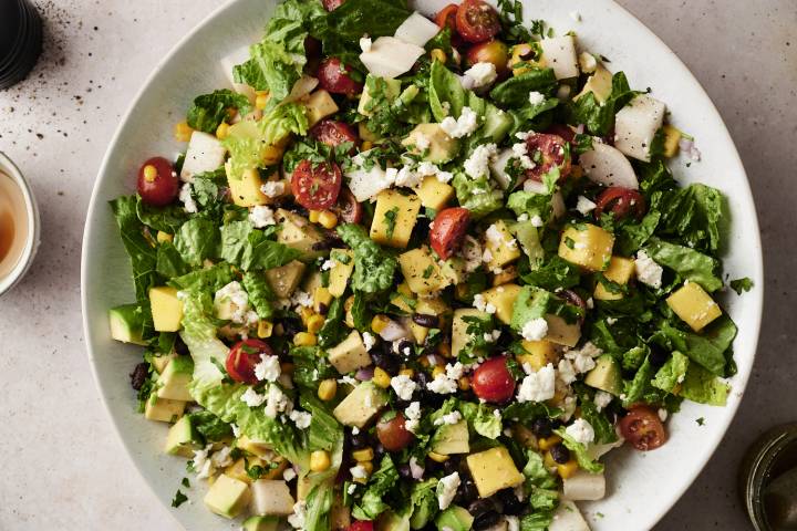 A vibrant Mexican chopped salad filled with diced mango, avocado, cherry tomatoes, black beans, corn, jicama, romaine lettuce, and crumbled cheese, garnished with fresh cilantro on a white plate.
