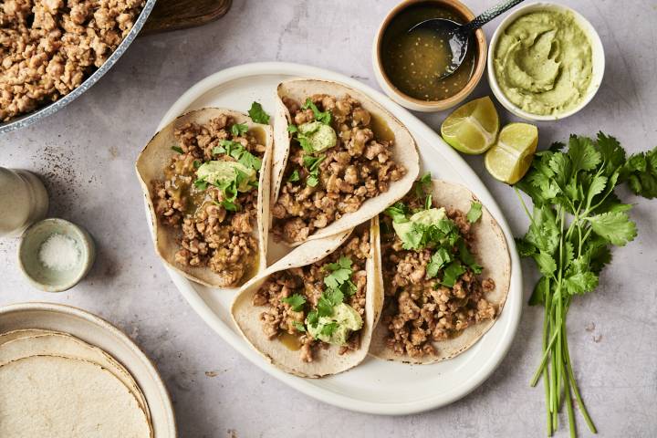 Plate of salsa verde turkey tacos with ground turkey filling, cilantro, and avocado crema, surrounded by lime wedges, salsa verde, and tortillas.