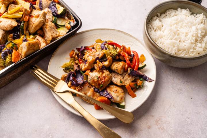 Stir-fried chicken with colorful vegetables, including red peppers and purple cabbage, on a white plate next to a bowl of white rice.