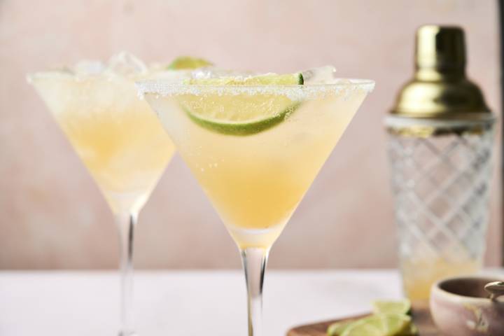 Two chilled cocktails with lime slices sit beside a cocktail shaker, set against a soft pink background.